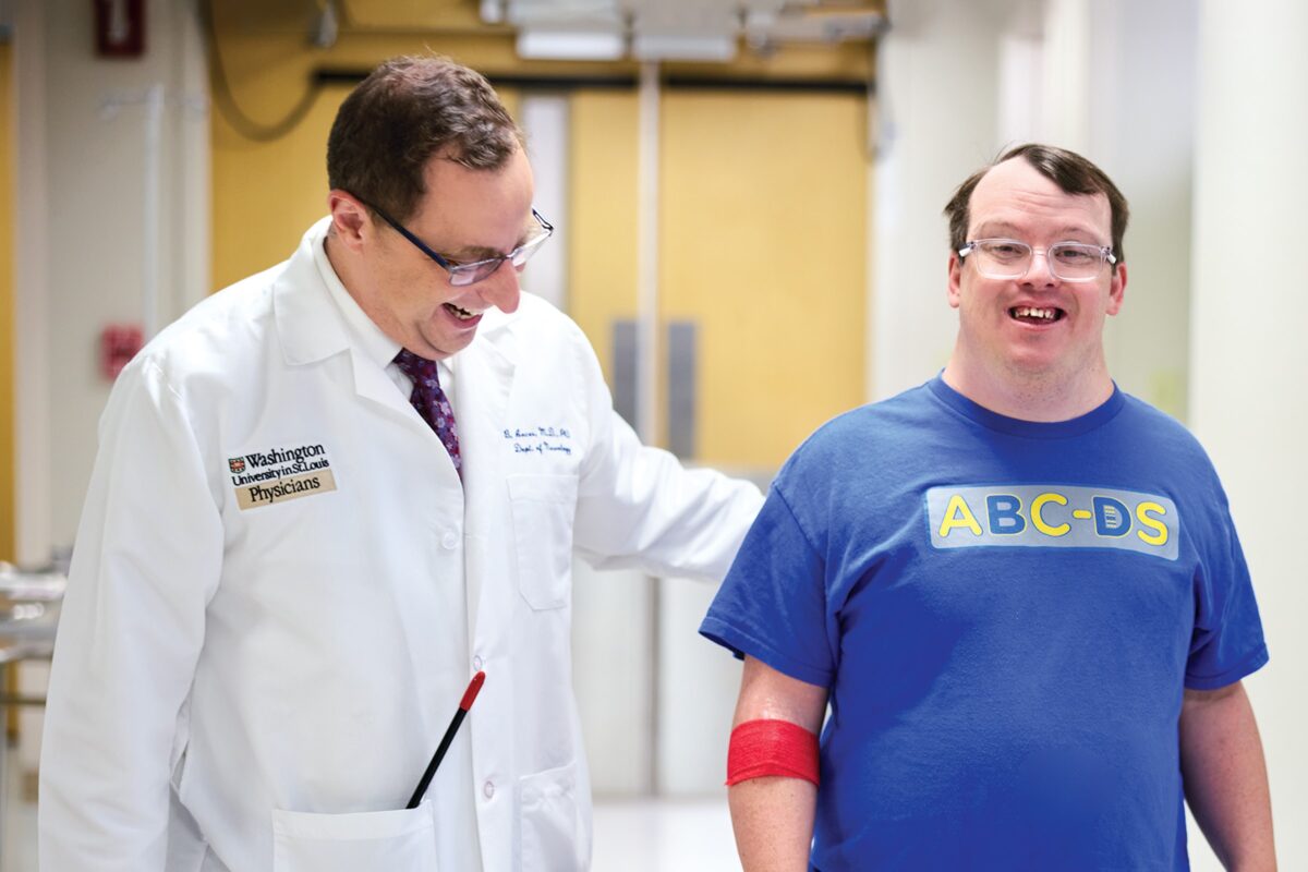 A smiling middle aged male doctor escorts a smiling male patient down the hallway of a hospital.